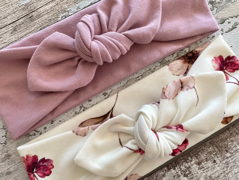 May include: Two fabric headbands, one is a solid dusty rose color and the other is white with a floral print. Both headbands are tied in a knot at the top.