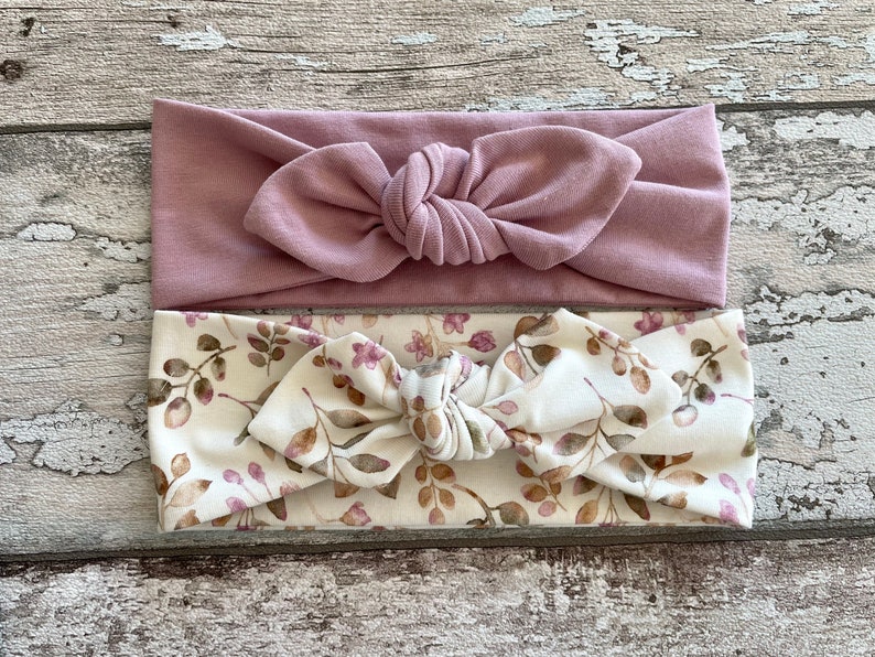 May include: Two headbands, one plain dusty rose and one with a floral pattern in shades of brown and pink on a white background.