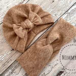 May include: A matching set of brown knitwear accessories, including a turban-style hat with a bow and a twisted headband. The items are displayed on a light-coloured wooden surface. The text "MOMMY&ME" is visible.