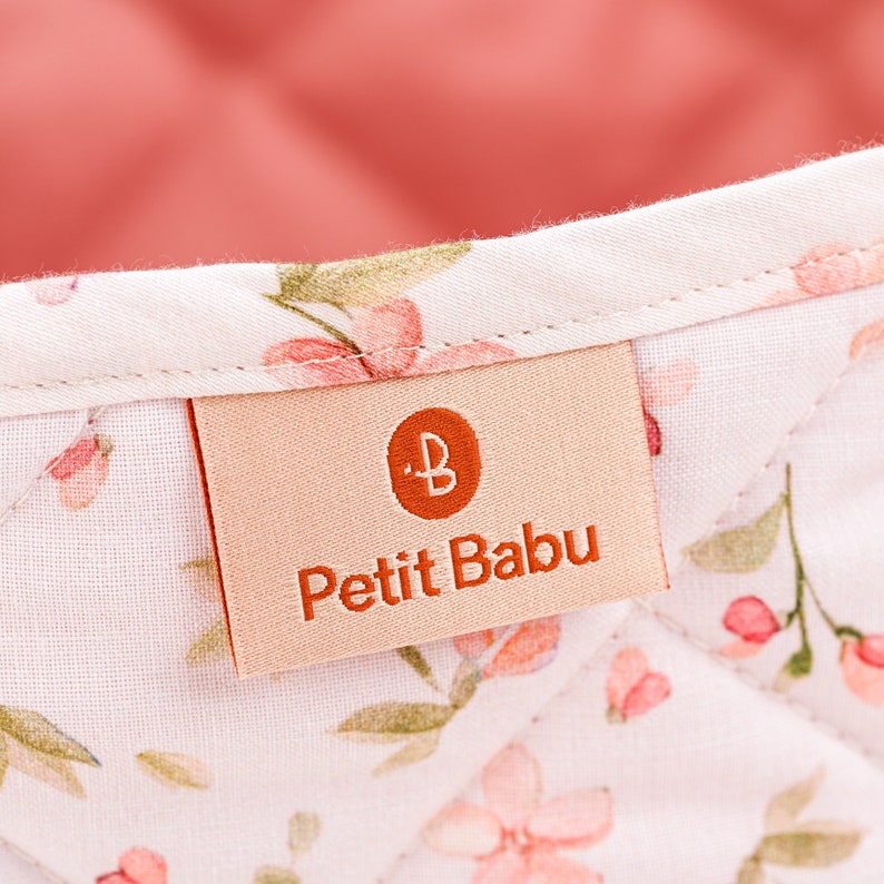 K&ouml;nnte beinhalten: Ein wei&szlig;er Stoffaufbewahrungskorb mit rosafarbenem Blumendruck und einem beigefarbenen Etikett mit der Aufschrift "Petit Babu" und einem kleinen orangen "B"-Logo.