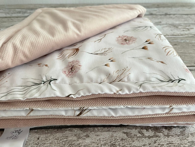 May include: A folded pink and white baby blanket with a floral pattern. The blanket is made of soft, plush fabric and is perfect for cuddling.