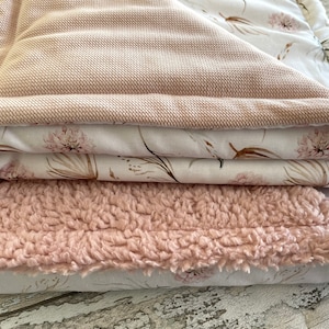 May include: A stack of three blankets in shades of pink and white. The top blanket is a soft, pink, textured fabric. The middle blanket is a white fabric with a delicate floral pattern in shades of pink and brown. The bottom blanket is a white fabric with a similar floral pattern.