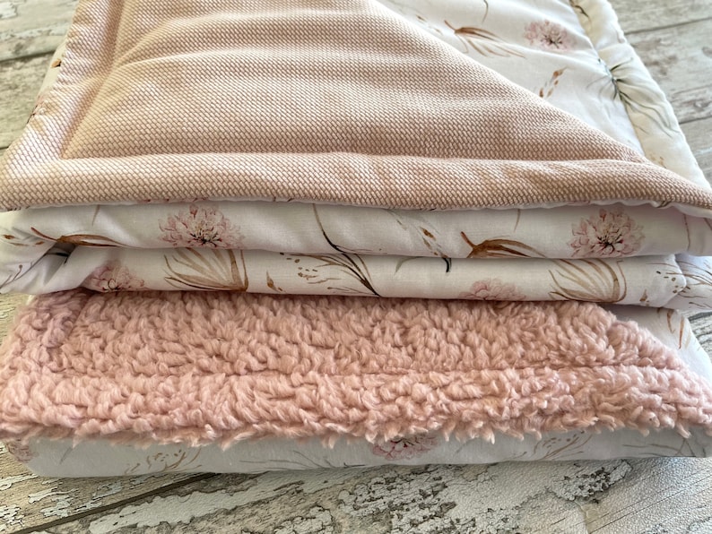 May include: A stack of three blankets in shades of pink and white. The top blanket is a soft, pink, textured fabric. The middle blanket is a white fabric with a delicate floral pattern in shades of pink and brown. The bottom blanket is a white fabric with a similar floral pattern.