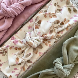 May include: A set of three knotted headbands. One is a solid dusty rose colour, one is a white headband with a brown and pink floral print, and the last is a solid sage green.