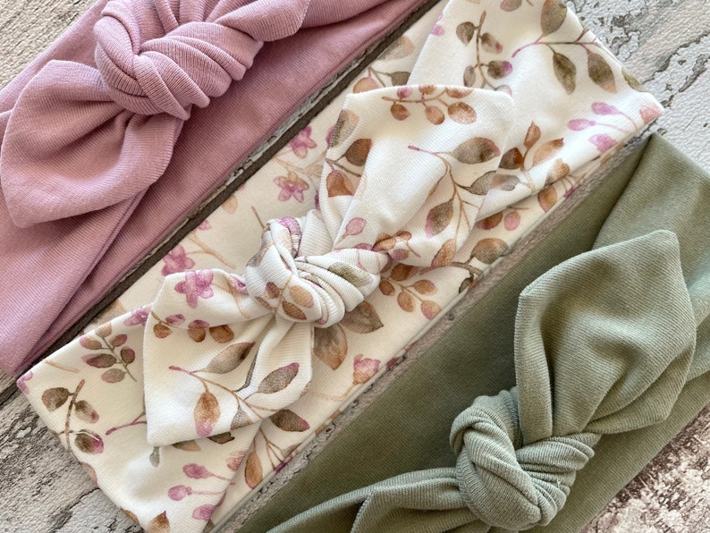 May include: A set of three knotted headbands. One is a solid dusty rose colour, one is a white headband with a brown and pink floral print, and the last is a solid sage green.