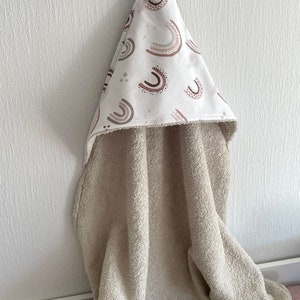 Peut inclure: Une serviette &agrave; capuche avec un corps en &eacute;ponge beige et une capuche en coton blanc. La capuche est orn&eacute;e d'un motif d'arcs-en-ciel et d'&eacute;toiles color&eacute;s.
