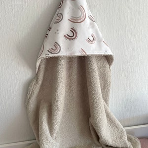 Peut inclure: Une serviette &agrave; capuche beige avec une capuche blanche orn&eacute;e d'un motif d'arcs-en-ciel bruns et roses et d'&eacute;toiles blanches.
