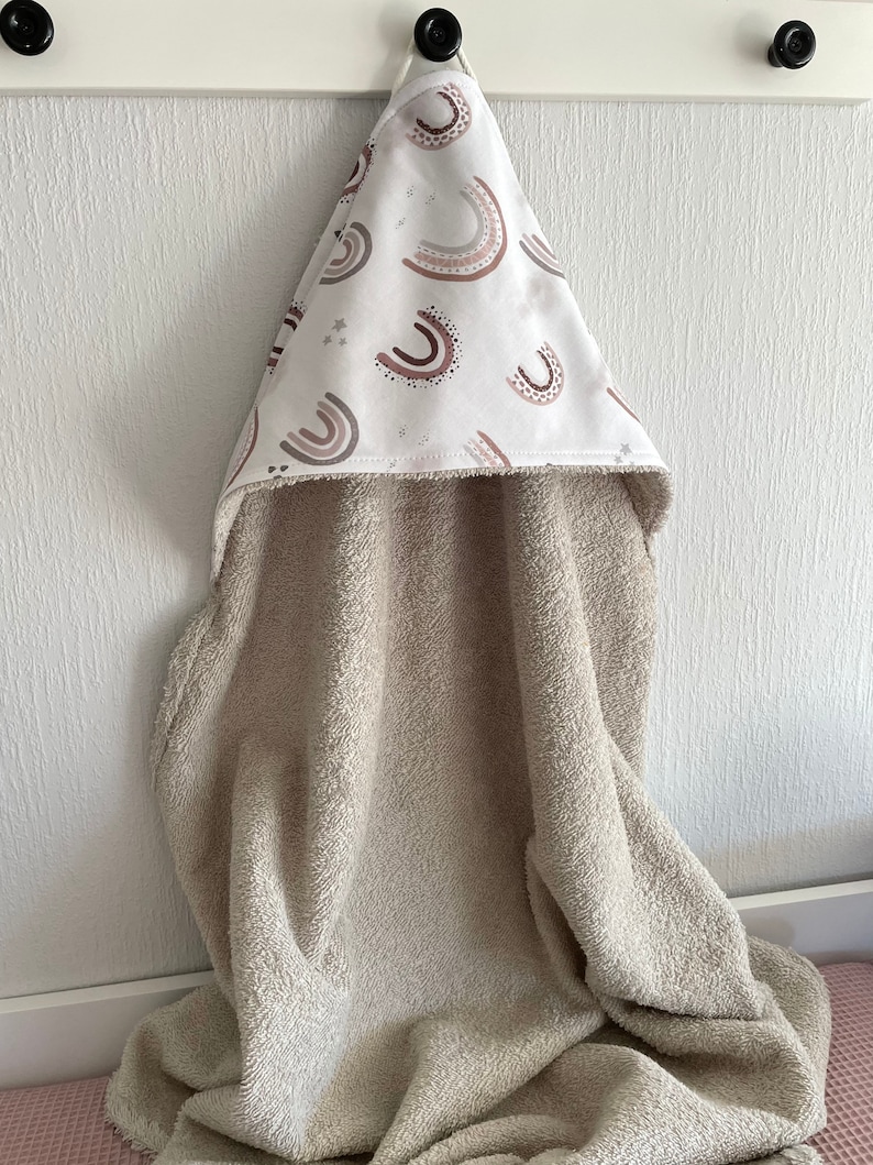 Peut inclure: Une serviette &agrave; capuche beige avec une capuche blanche orn&eacute;e d'un motif d'arcs-en-ciel bruns et roses et d'&eacute;toiles blanches.