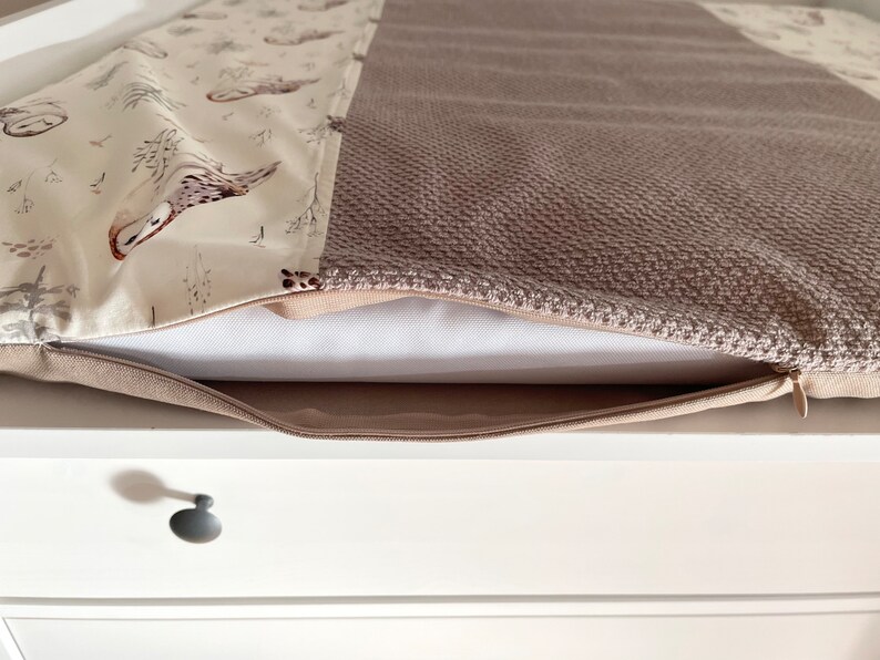 May include: A beige and brown pillow cover with a zipper closure. One side features a pattern of owls and trees on a cream background. The other side has a textured, woven appearance in a solid brown color. The pillow is resting on a white surface.