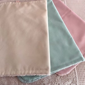 K&ouml;nnte beinhalten: Drei Stoffquadrate mit Fransen. Die Quadrate sind beige, hellgr&uuml;n und hellrosa.