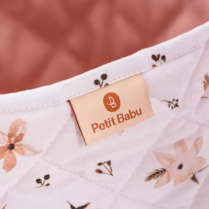 K&ouml;nnte beinhalten: Ein wei&szlig;er gesteppter Aufbewahrungskorb mit rosa Futter und braunem Blumendruck. Der Korb hat ein Etikett mit der Aufschrift "Petit Babu" und einem kleinen "B"-Logo.