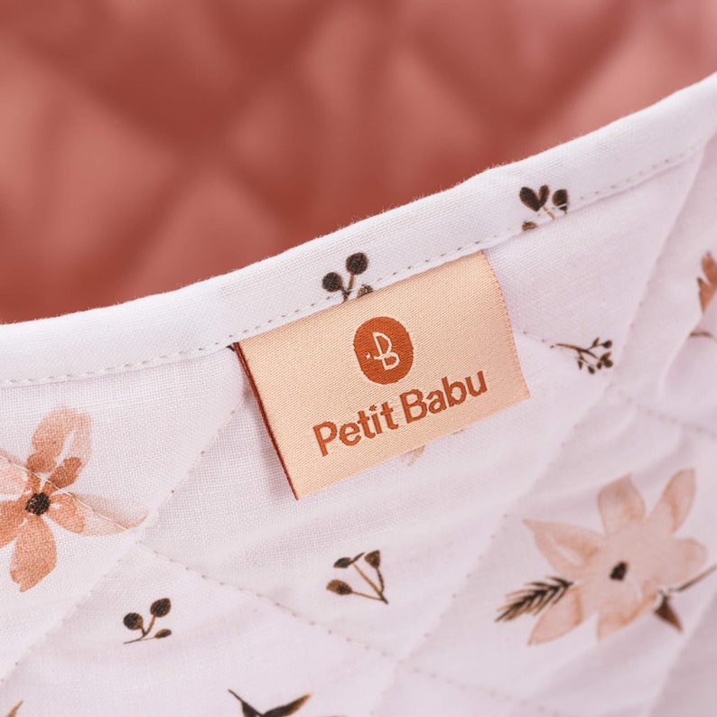 K&ouml;nnte beinhalten: Ein wei&szlig;er gesteppter Aufbewahrungskorb mit rosa Futter und braunem Blumendruck. Der Korb hat ein Etikett mit der Aufschrift "Petit Babu" und einem kleinen "B"-Logo.