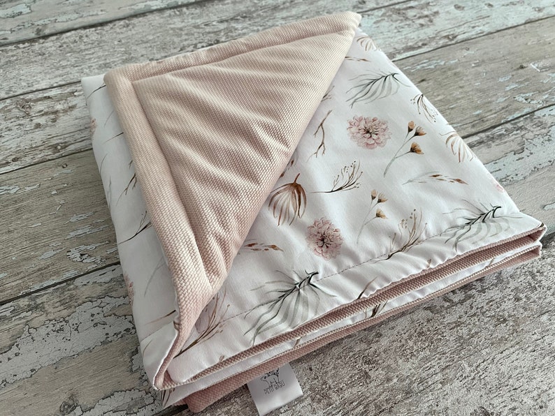 May include: A soft, pink and white baby blanket with a floral pattern. The blanket is folded in half and has a pink velvet backing.