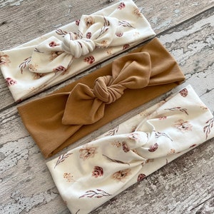 May include: Three fabric headbands with a knotted bow design. Two headbands feature a white background with a floral pattern in shades of brown and pink. The third headband is solid brown.