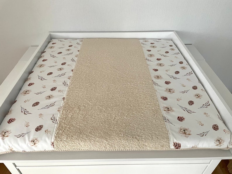 May include: A white changing pad cover with a beige, fluffy, textured center and two side panels with a white background and a brown floral pattern.