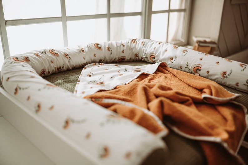 May include: A white and orange baby nest with a rainbow pattern and a brown blanket inside.