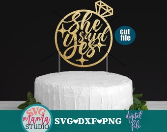 Puede incluir: Topper de pastel de purpurina dorada con el texto "She said yes" dentro de un círculo con un anillo de diamantes en la parte superior. El topper está en un pastel blanco con follaje verde en el fondo.