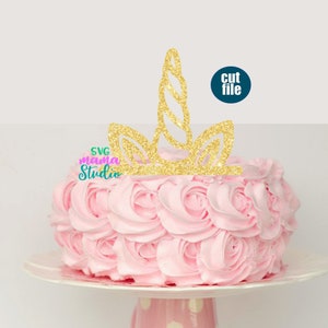 May include: Gold glitter unicorn cake topper with a curled horn and ears. The topper is on top of a pink frosted cake.