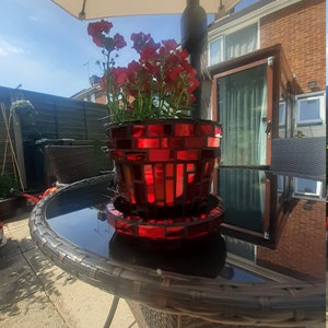 May include: A red mosaic glass plant pot, filled with red flowers, sits on a matching saucer. The pot is on a dark glass table with a woven edge, under a white parasol. The scene is outdoors on a sunny day.