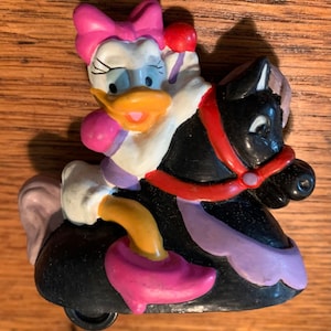 May include: A colorful figurine of Daisy Duck riding a black horse. Daisy Duck is wearing a pink bow, gloves, and shoes. The horse has a red bridle and purple saddle. The figurine is on a wooden surface.