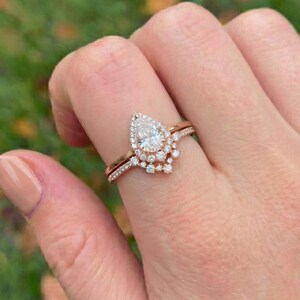May include: A rose gold engagement ring set featuring a pear-shaped center stone surrounded by a halo of small round diamonds, with a matching pavé diamond band nestled against the main setting, creating a layered and sparkling appearance.