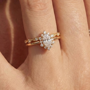 May include: A gold engagement ring featuring an oval diamond surrounded by smaller stones. A second gold band with a decorative pattern sits beneath. The rings are displayed on a finger, with a soft-focus background.