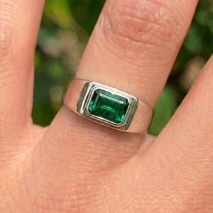 May include: A silver-toned ring featuring a rectangular, emerald-cut green gemstone set in a smooth, polished metal bezel. The band has a wide, flat profile that tapers slightly toward the sides, showcasing a clean and solid construction.