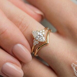 Puede incluir: Un conjunto de anillos de compromiso de oro con un diamante central de talla marquesa flanqueado por tres pequeños diamantes redondos a cada lado, junto a una alianza de boda de oro a juego con forma de chevrón.