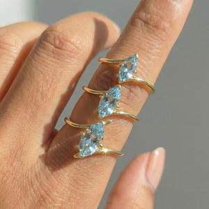 May include: Three gold rings with marquise-cut, light blue gemstones are displayed on a finger. The rings have a delicate, minimalist design, with the gemstone held by thin gold bands. The background is a soft, neutral tone.
