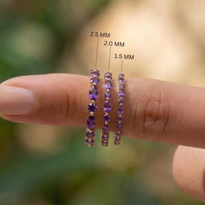 May include: Three gold rings with purple gemstones of varying sizes are displayed on a finger. The rings are labeled 2.5 MM, 2.0 MM, and 1.5 MM. The gemstones are oval and round, set in a gold band.