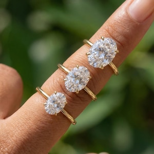 May include: Three gold rings with oval-cut diamonds are displayed on a finger. The rings have a classic, minimalist design with a thin gold band. The diamonds sparkle brightly against a blurred green background.