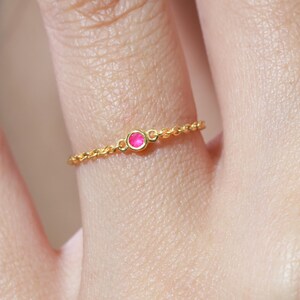 Könnte beinhalten: Ein zarter Ring aus einer goldenen Kette mit einem einzelnen, kleinen, runden, leuchtend pinkfarbenen Edelstein, der in einer runden goldenen Fassung sitzt. Der Ring besteht aus einer feinen, gedrehten Goldkettengliederung, die den Finger umschließt.