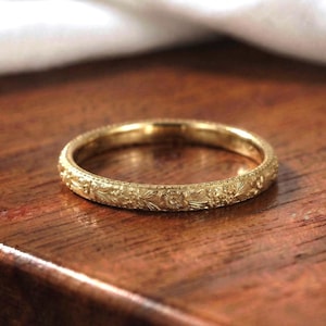 Könnte beinhalten: Ein goldener Ring mit einer durchgehenden, komplizierten Blumengravur, die sich um die gesamte Außenfläche zieht. Das Metall hat eine polierte Oberfläche, die die detaillierten botanischen Muster hervorhebt, welche in das kreisförmige Schmuckstück eingraviert sind.