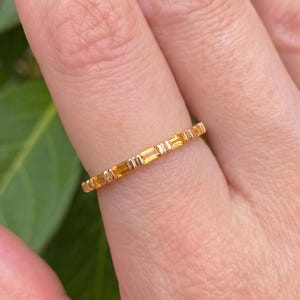 Op de afbeelding: Een dunne gouden ring met een reeks afwisselende gele edelstenen in baguette-slijpvorm en kleine gouden metalen staafjes. De ring heeft een gepolijste afwerking en een slank profiel, dat de vinger omsluit met een herhalend patroon van stenen.