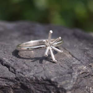 May include: A silver-colored metal ring featuring a six-prong setting designed to hold a gemstone. The band is smooth and polished, and the prongs extend upward from the center of the ring, creating a star-like structure for a stone setting.