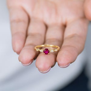 May include: A gold ring featuring a round, vibrant red gemstone at the center, flanked by two small, marquise-shaped clear stones on each side of the band. The thin, polished gold band holds the central red stone in a four-prong setting.