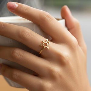May include: A gold-toned ring featuring a chemical molecular structure design on the band. The ring is worn on a finger, positioned against a white mug containing a hot beverage with visible steam rising from the top of the cup.