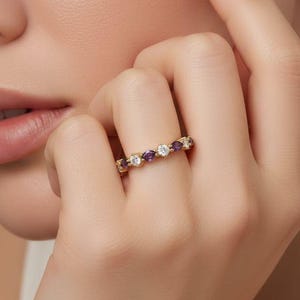 May include: A gold band ring featuring an alternating pattern of round clear stones and marquise-cut purple stones. The ring is worn on a finger, showcasing the continuous arrangement of gemstones set within the metallic band structure.