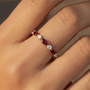 Puede incluir: Un anillo de banda dorada con un patrón alterno de rubíes rojo oscuro en talla marquesa y diamantes blancos transparentes en talla redonda. Las piedras están engastadas en fila a lo largo de la fina banda metálica, creando un diseño simétrico.