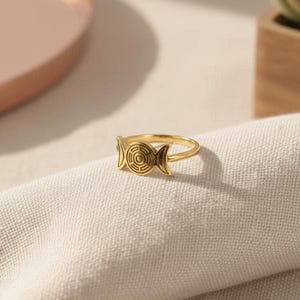May include: A gold-toned metal ring featuring a central circular emblem with concentric lines flanked by two crescent moon shapes on either side. The band is thin and polished, showcasing a smooth metallic finish throughout the entire jewelry piece.
