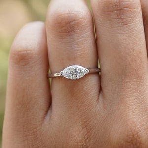 May include: A silver ring with a marquise-cut diamond center stone. The diamond is set horizontally, and the band is thin and polished. The ring is shown on a person's finger against a blurred background.