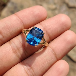 May include: A gold ring with a large, rectangular, bright blue gemstone. The gemstone is held in place by four prongs, and the band has a split design. The ring is shown on a hand.
