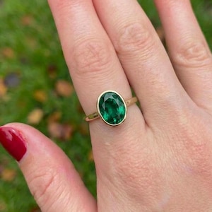 May include: A gold ring featuring a single oval-shaped green gemstone set in a smooth bezel setting. The band is thin and polished, showcasing a minimalist design that highlights the vibrant green color of the central stone against the gold metal.