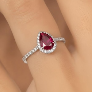 Könnte beinhalten: Ein silberfarbener Ring mit einem birnenförmigen, tiefroten Edelstein in der Mitte. Der zentrale Stein ist von einem Kranz aus kleinen, klaren, rund geschliffenen Steinen umgeben. Die Ringschiene ist schmal und mit einer Reihe kleiner Steine besetzt.