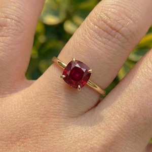 Pode incluir: Um anel de ouro com uma única pedra preciosa vermelho-escura em corte almofada, montada em quatro garras. A banda é de metal dourado simples e polido. A pedra preciosa está centrada no anel, exibindo uma cor vermelha vibrante e saturada.