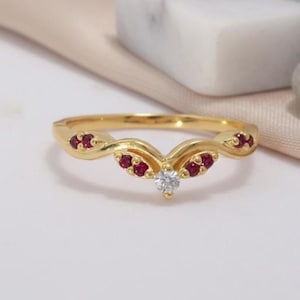 May include: A gold-colored ring with a curved design. The ring features small red gemstones and a single, larger clear gemstone at the center. The ring is set against a neutral background.