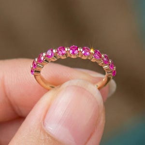May include: A gold band ring featuring a row of eleven round, vibrant pink gemstones set in a shared prong setting. The metal has a polished finish, and the stones are arranged in a continuous arc across the top half of the circular band.