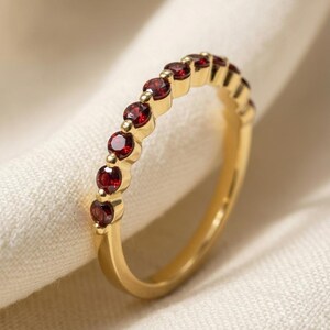 Op de afbeelding: Een gouden ring met een rij kleine, ronde, dieprode granaatedelstenen in een klauwzetting. De ring heeft een gepolijste metalen afwerking en volgt een klassiek eternity-ontwerp waarbij de stenen langs de bovenste helft zijn uitgelijnd.