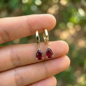 May include: A pair of gold hoop earrings featuring dangling teardrop-shaped deep red gemstones. Each earring consists of a polished gold hoop connected to a single faceted pear-cut red stone, creating a simple and elegant jewelry design.