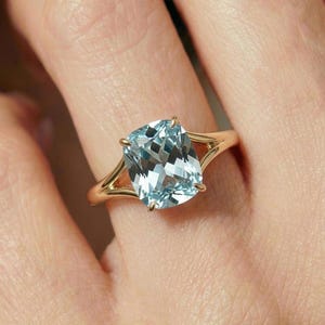 May include: A gold ring featuring a large, cushion-cut, light blue aquamarine gemstone held by four prongs. The band is crafted from polished yellow gold and features a split-shank design that tapers as it meets the central gemstone setting.